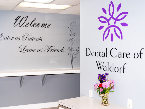 Reception area wall featuring a "Welcome, Enter as Patients, Leave as Friends" sign beside the Dental Care of Waldorf logo and a vase of colorful flowers