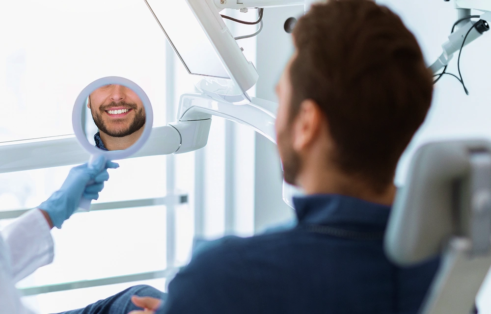 Man seated in a dental treatment chair with his smile reflected in a mirror